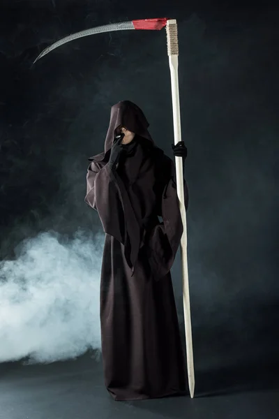 Full length view of woman in death costume holding scythe and smoking cigarette on black — Stock Photo