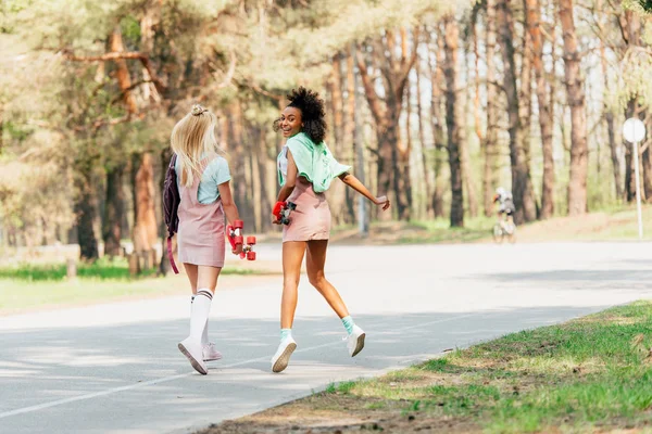 Full length view of laughing multiethnic friends with penny boards jumping on road — Stock Photo