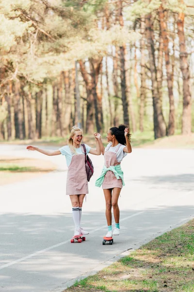 Vista completa de dos sonrientes amigos multiculturales tomados de la mano mientras patinan en penny boards en la carretera - foto de stock