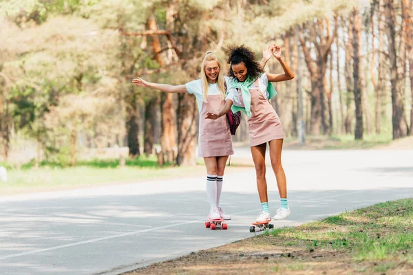 Full length view of two smiling multicultural friends skateboarding on penny boards on road — Stock Photo