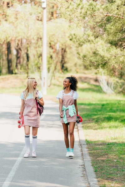 Ganzkörperansicht von zwei lächelnden multikulturellen Freunden mit Penny-Tafeln, die einander anschauen und auf der Straße gehen — Stockfoto