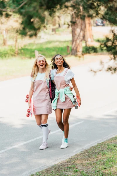 Full length view of two multicultural friends with penny boards embracing while walking on road — Stock Photo