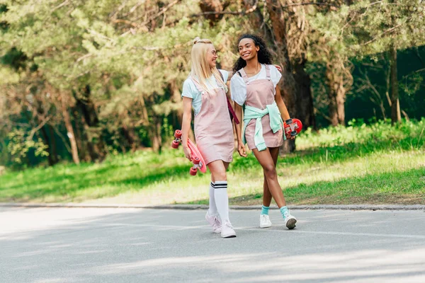 Vue pleine longueur de deux amis multiculturels avec des planches de penny embrassant tout en marchant sur la route — Photo de stock