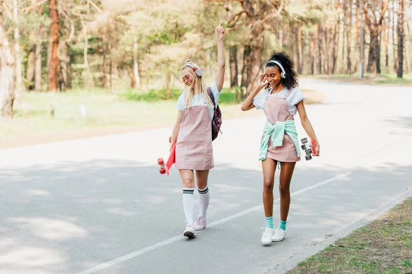 Full length view of two smiling multicultural friends with penny boards walking on road and listening music in headphones — Stock Photo