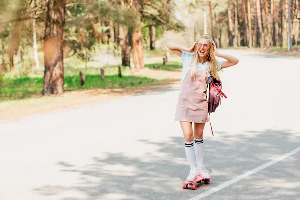 Vue complète de fille blonde excitée skateboard et écouter de la musique dans les écouteurs — Photo de stock