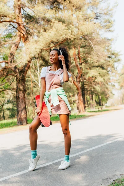 Vue complète de la fille afro-américaine dans les écouteurs debout sur la route et tenant penny conseil — Photo de stock