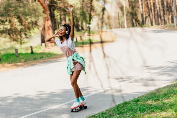 Vista completa de la chica afroamericana skateboarding y escuchar música en auriculares - foto de stock