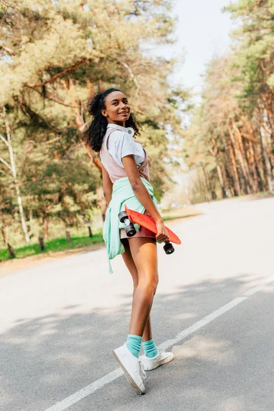 Full length view of african american girl in headphones standing on road and holding penny board — Stock Photo