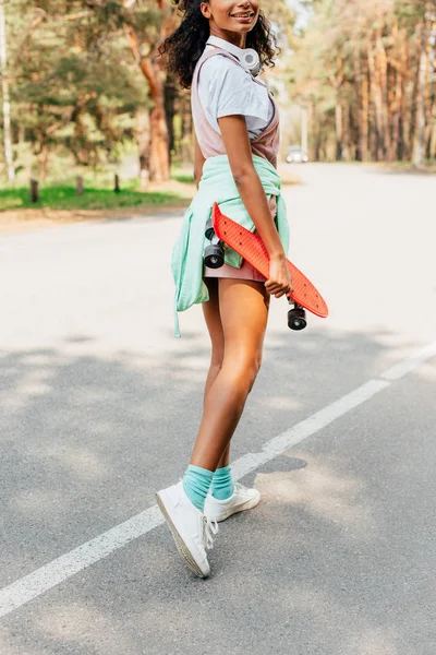 Vista recortada de chica afroamericana de pie en la carretera y la celebración de penny board - foto de stock