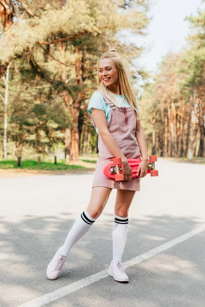 Full length view of smiling blonde girl standing on road and holding penny board — Stock Photo