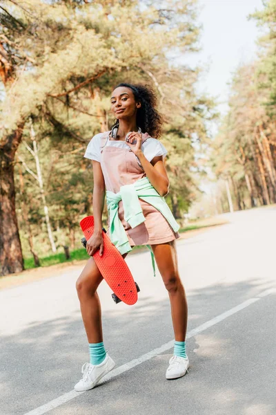 Full length view of african american girl in headphones standing on road and holding penny board — Stock Photo