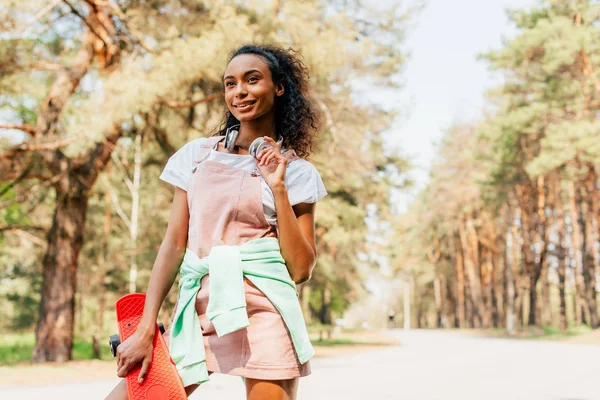 African american girl in headphones standing on road and holding penny board — Stock Photo