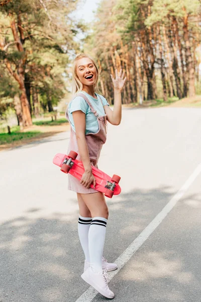 Full length view of smiling blonde girl standing on road with penny board and waving hand — Stock Photo