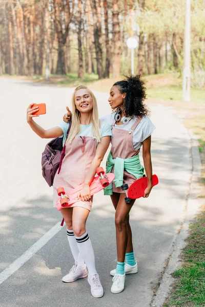 Vue pleine longueur de deux amis multiculturels souriants avec des planches de penny prendre selfie sur la route — Photo de stock