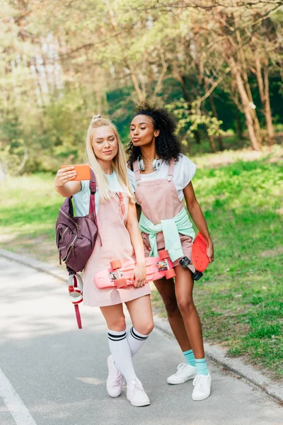 Vue pleine longueur de deux amis multiculturels souriants avec des planches de penny prendre selfie sur la route — Photo de stock