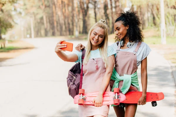 Dos sonrientes amigos multiétnicos sosteniendo tableros de peniques y tomando selfie en la carretera - foto de stock