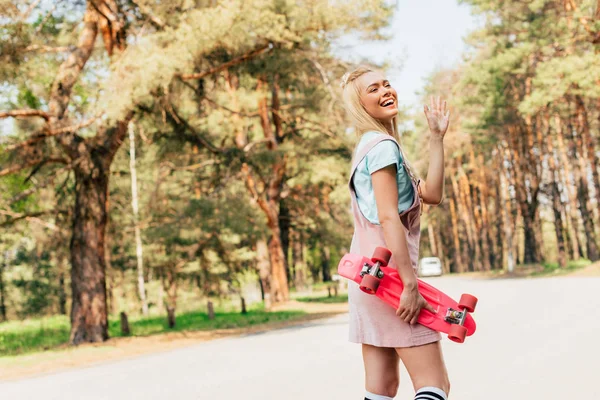 Excited blonde girl holding penny board and waving hand on road — Stock Photo