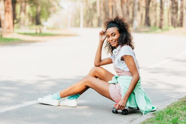 Sorrindo menina americana africana com fones de ouvido sentado na placa penny na estrada — Fotografia de Stock