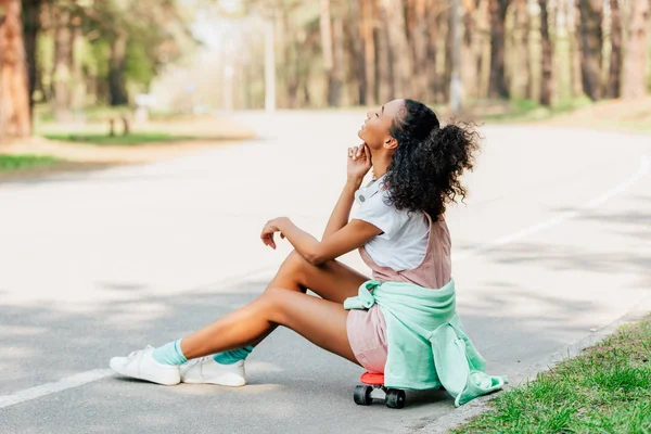 Smiling african american girl with headphones sitting on penny board on road — Stock Photo