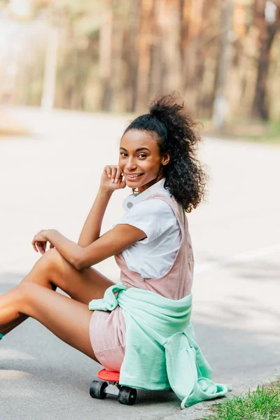 Sonriente afroamericana chica con auriculares sentado en penny board en la carretera - foto de stock