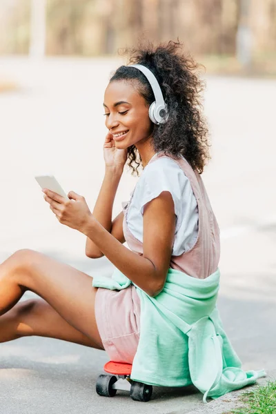 Smiling african american girl listening music in headphones and using smartphone while sitting on penny board — Stock Photo