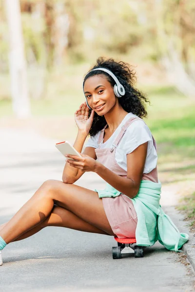 Улыбающаяся африканская американка, слушающая музыку в наушниках и использующая смартфон, сидя на доске — стоковое фото
