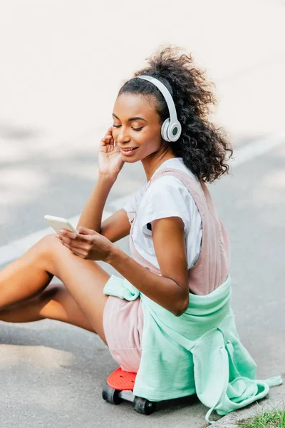 Smiling african american girl listening music in headphones and using smartphone while sitting on penny board — Stock Photo