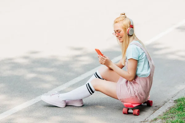 Sonriente chica rubia escuchando música en los auriculares mientras está sentado en penny board en la carretera - foto de stock