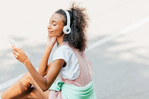 Chica afroamericana sonriente escuchando música en los auriculares y usando el teléfono inteligente en la carretera - foto de stock