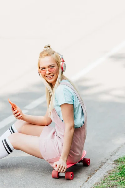 Lächelndes blondes Mädchen, das Musik mit Kopfhörern hört, während es auf dem Penny Board unterwegs sitzt — Stockfoto