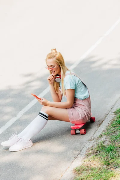 Souriant fille blonde écouter de la musique dans les écouteurs tout en étant assis sur le panneau penny sur la route — Photo de stock