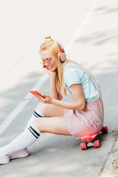 Smiling blonde girl listening music in headphones while sitting on skateboard on road — Stock Photo