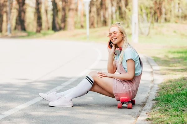 Sonriente chica rubia en gafas de sol sentado en penny board y hablando en el teléfono inteligente en la carretera - foto de stock