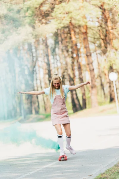 Vue pleine longueur de fille blonde agitant les mains tout en skateboard dans la fumée verte sur la route — Photo de stock