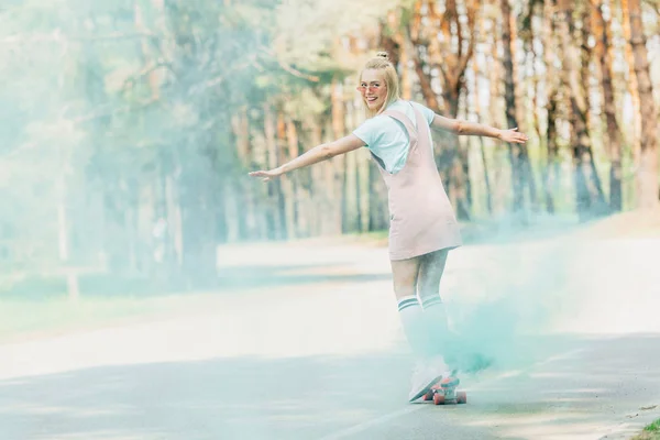 Full length view of smiling blonde girl waving hands while skateboarding in green smoke on road — Stock Photo