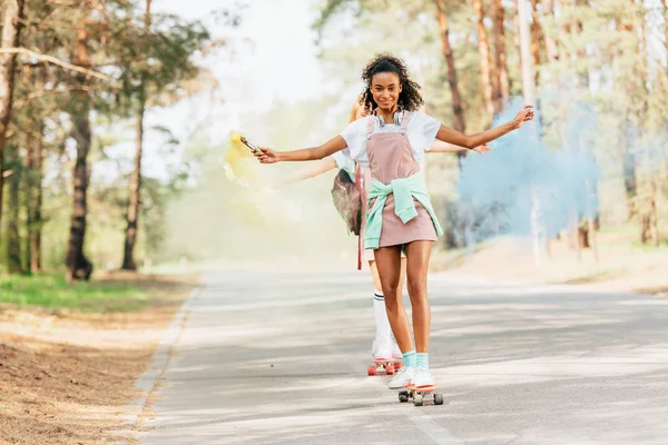Full length view of two girls skateboarding with blue and yellow smoke grenade on road — Stock Photo