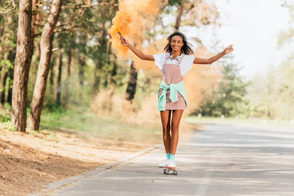 Pleine longueur vue de sourire afro-américaine fille skateboard et tenant grenade fumigène orange sur la route — Photo de stock