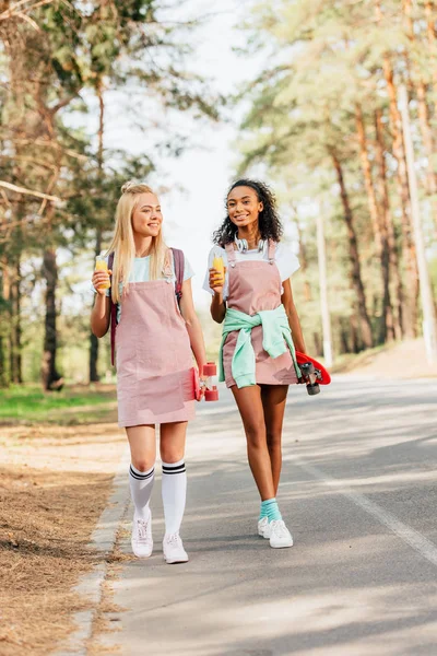 Vista completa de dos amigos multiculturales con tableros de peniques sosteniendo botellas de jugo de naranja mientras camina por la carretera - foto de stock