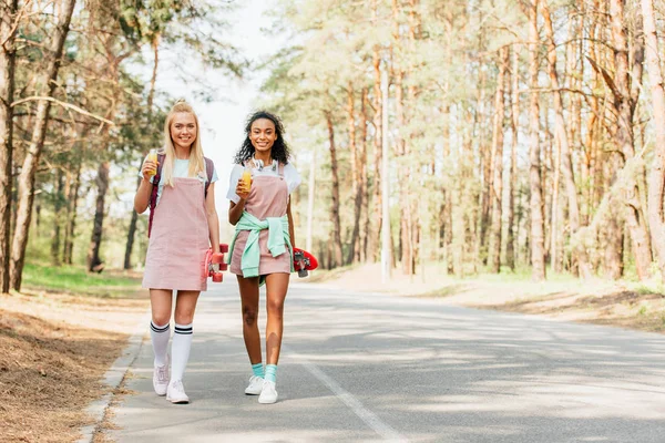 Full length view of two multicultural friends with penny boards holding bottles of orange juice while walking on road — Stock Photo