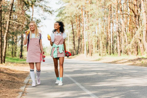 Full length view of two multiethnic friends with penny boards holding bottles of orange juice on road — Stock Photo