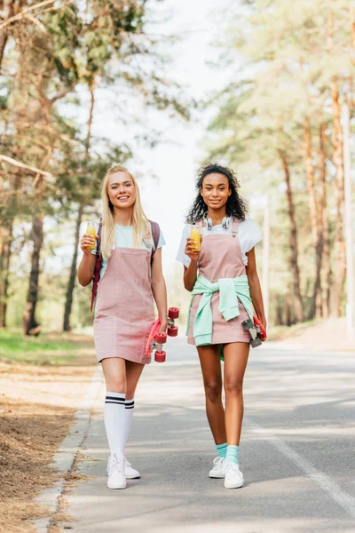 Full length view of two multiethnic friends with penny boards holding bottles of orange juice on road — Stock Photo