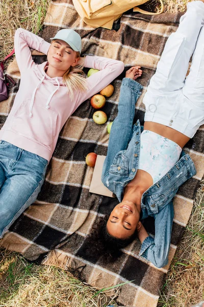 Вид сверху на двух улыбающихся мультиэтнических девушек лежащих на клетчатом одеяле с яблоками — стоковое фото