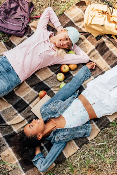 Vue de dessus de deux filles multiethniques souriantes allongées sur une couverture à carreaux avec des pommes — Photo de stock