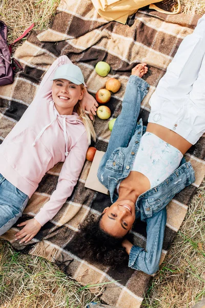 Vista superior de duas meninas multiétnicas sorridentes deitadas no cobertor xadrez com maçãs — Fotografia de Stock