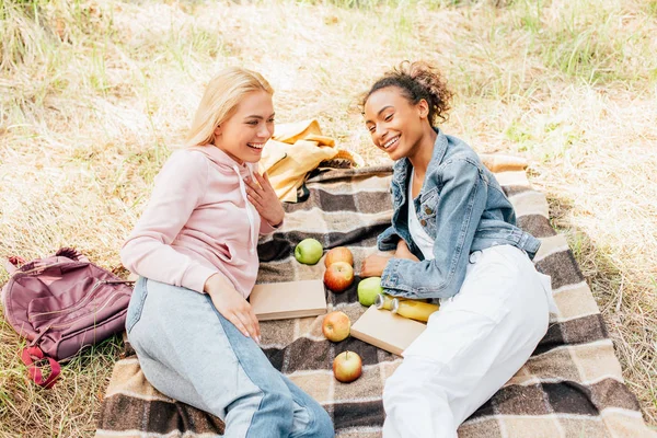 Dos amigos multiétnicos sonrientes en manta a cuadros con manzanas - foto de stock