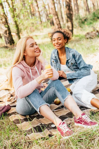 Zwei multiethnische Mädchen sitzen auf karierten Decken und halten Flaschen Orangensaft in der Hand — Stockfoto