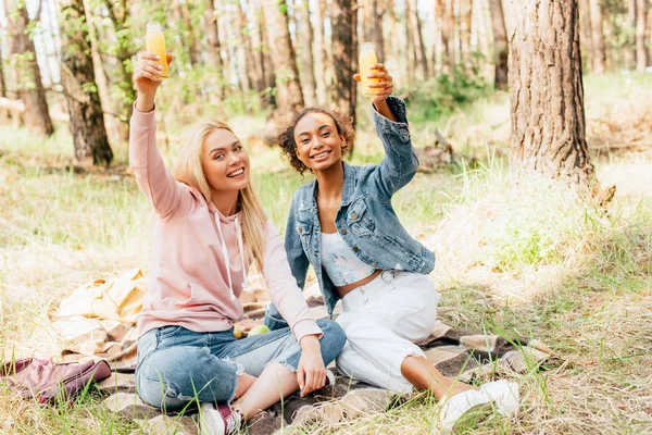 Duas meninas multiétnicas sentadas no cobertor xadrez e levantando garrafas de suco de laranja — Fotografia de Stock