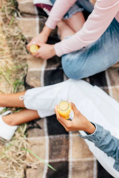 Vista recortada de dos niñas sosteniendo botellas en jugo de naranja mientras se sienta en manta a cuadros en el suelo - foto de stock