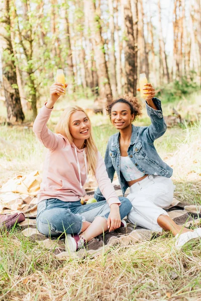 Dos chicas multiétnicas sentadas en manta a cuadros y criando botellas de jugo de naranja - foto de stock