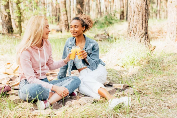 Dos chicas multiétnicas sentadas en manta a cuadros y sosteniendo botellas de jugo de naranja - foto de stock